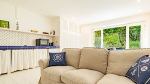 A living room with a beige sofa, a blue cushion with a compass design, a kitchen area with a microwave, and a window with a view of greenery.