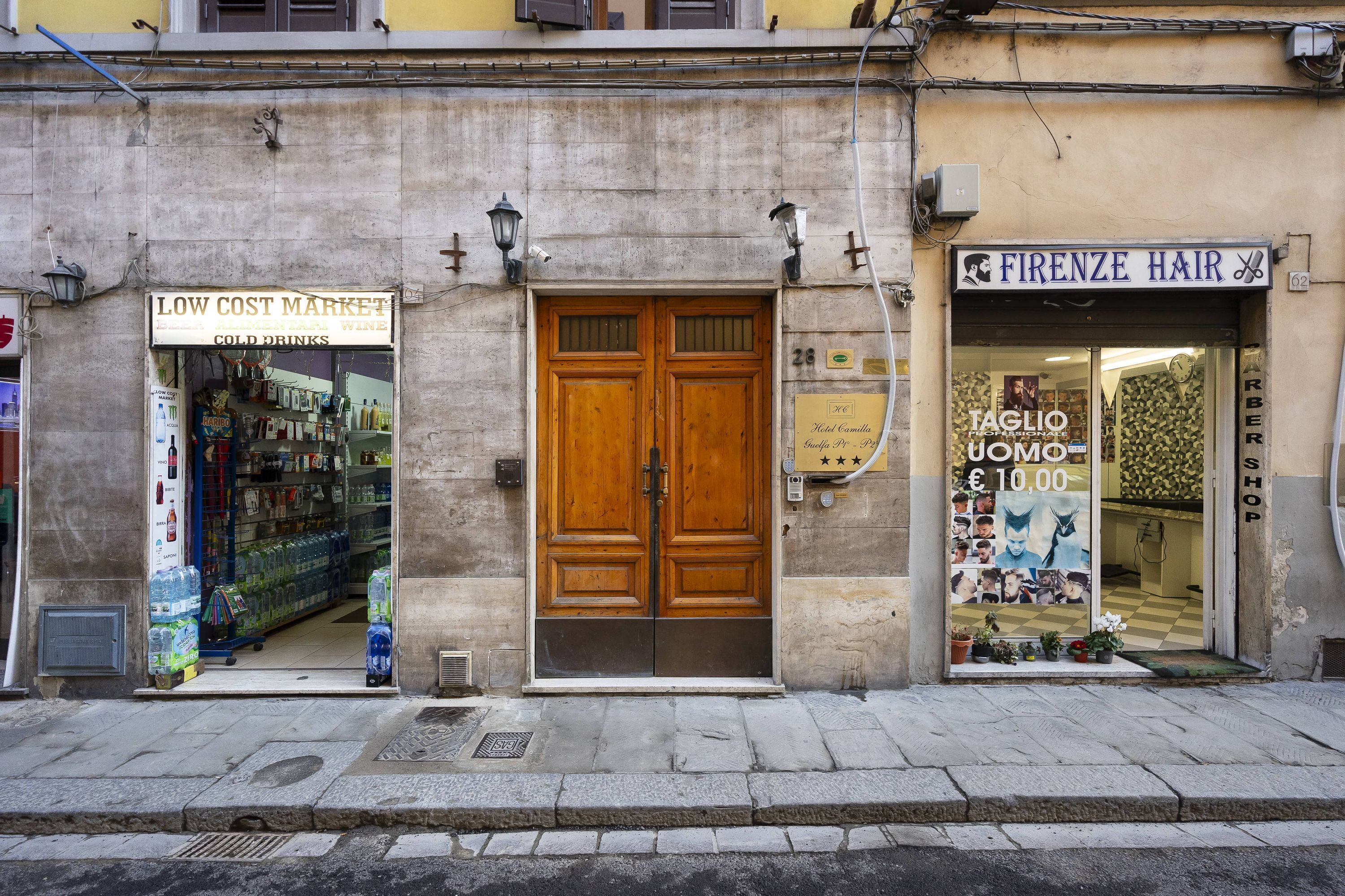 Une devanture de magasin avec une porte en bois, une enseigne indiquant « FIRENZE HAIR » et une autre, « LOW COST MARKET », avec de l’eau en bouteille et des en-cas exposés à l’intérieur.