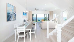 A spacious living area with a dining table, white furniture, and a view of the ocean through large windows. - Oceanfront 3BR + Private Dock at Sea Haven – Cassava Point Villa 4D (Patricks Island)