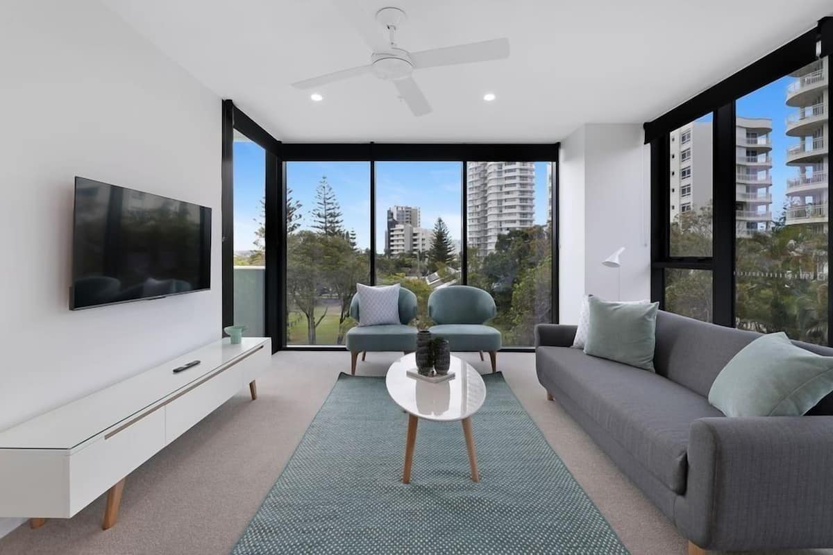 A modern living room with a flat-screen TV, a sofa, a coffee table, and a large window offering a view of tall buildings and trees.