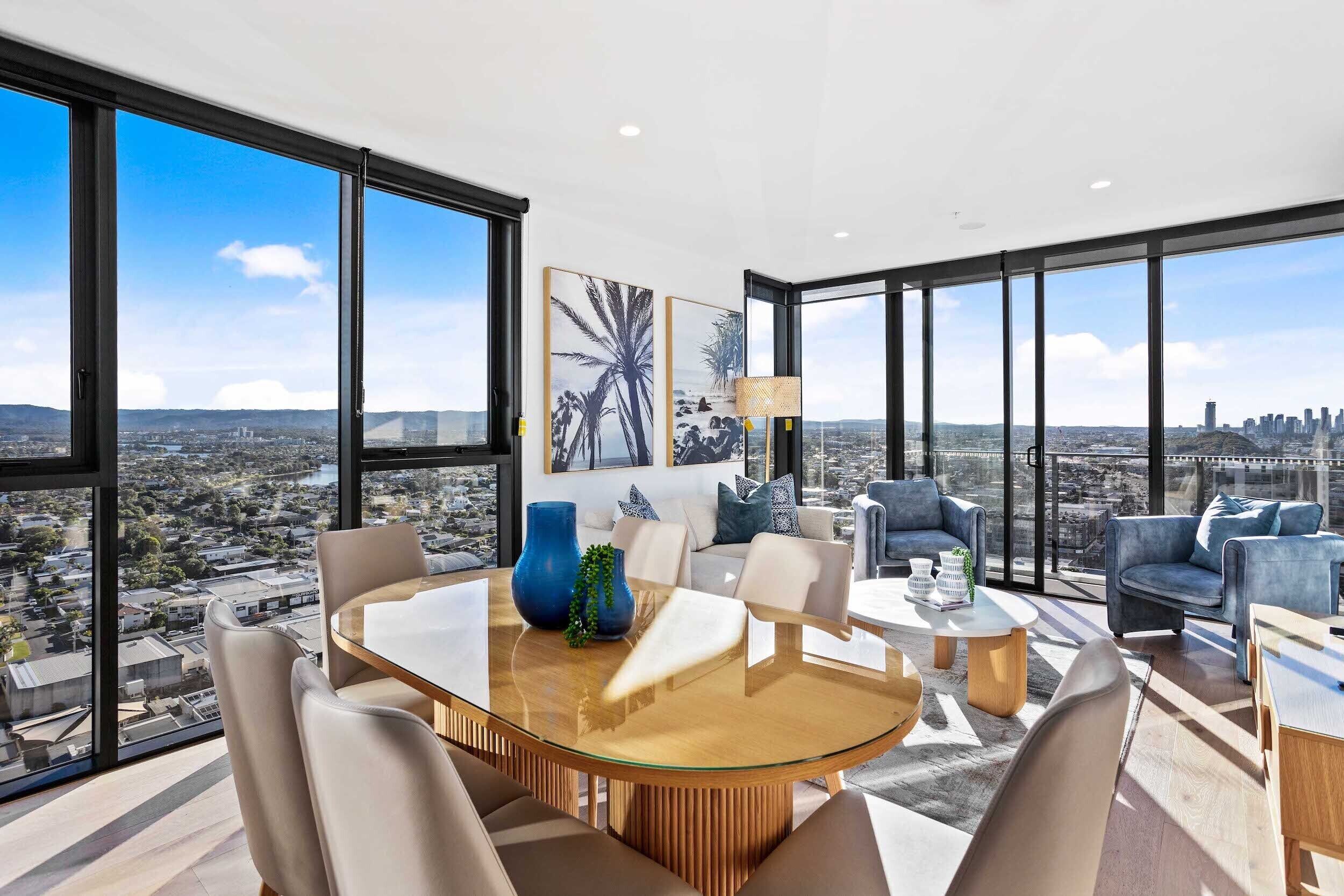 A modern living room with a glass table, comfortable seating, and large windows offering a city view.