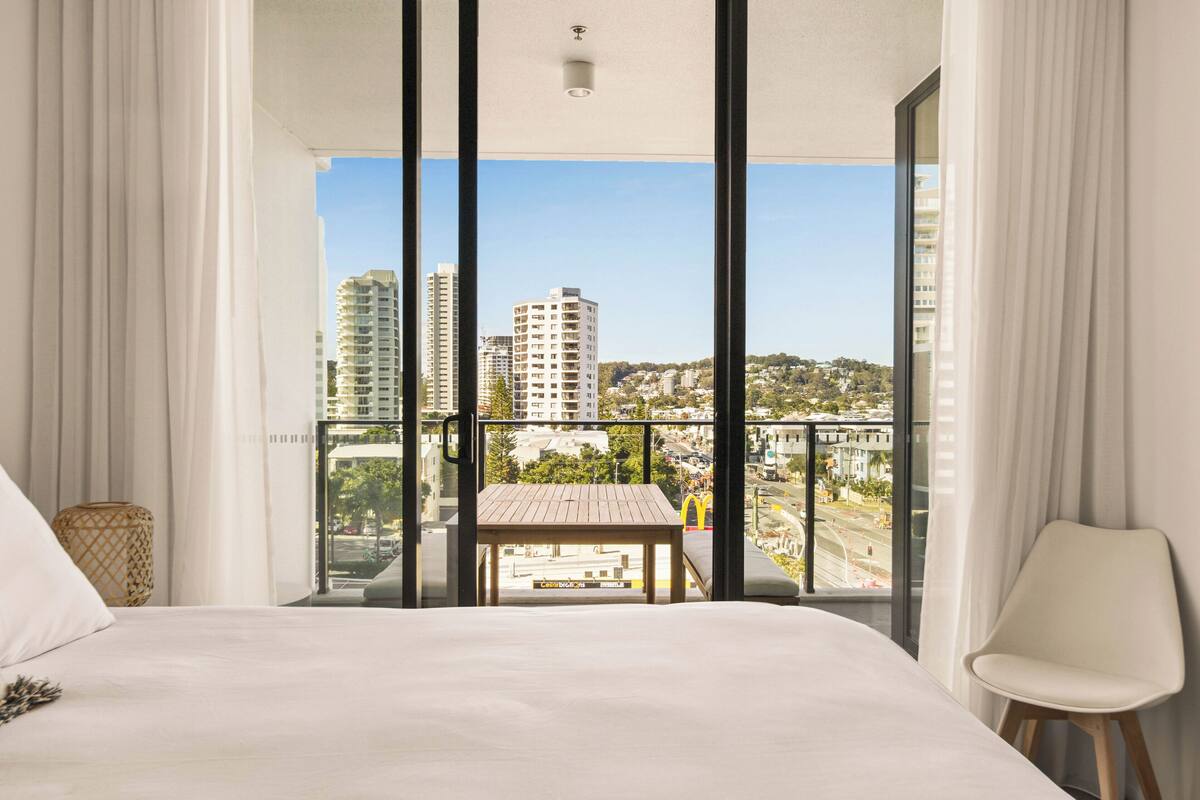 A hotel room with a large bed, a view of the city through floor-to-ceiling windows, and a balcony with a table and chair.