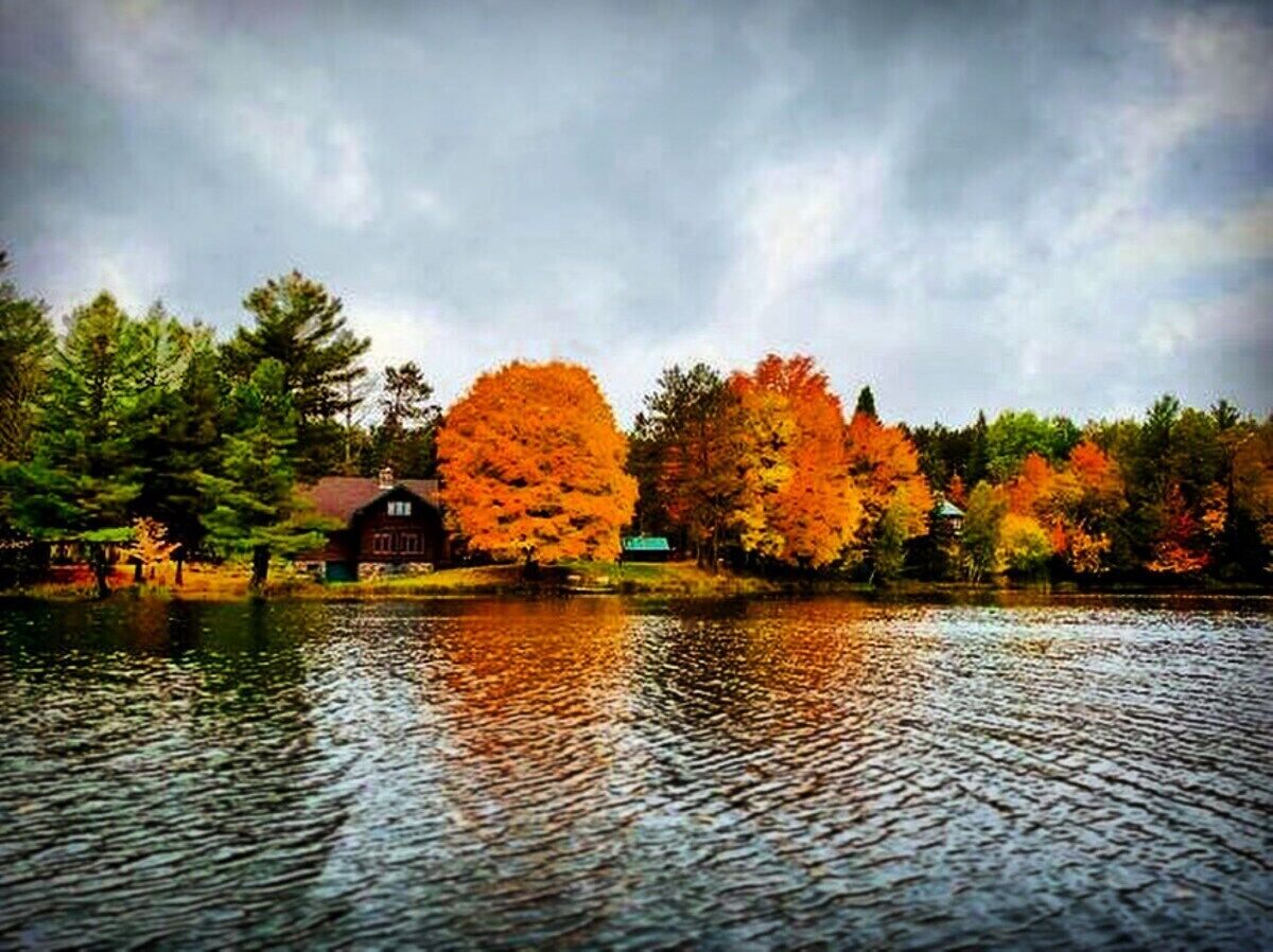 A lake with autumn trees reflecting on the water, a red building, and a small structure in the background.