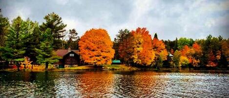 A lake with autumn trees reflecting on the water, a red building, and a small structure in the background.