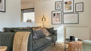A cozy living room with a grey sofa, a woven ottoman, and a floor lamp.