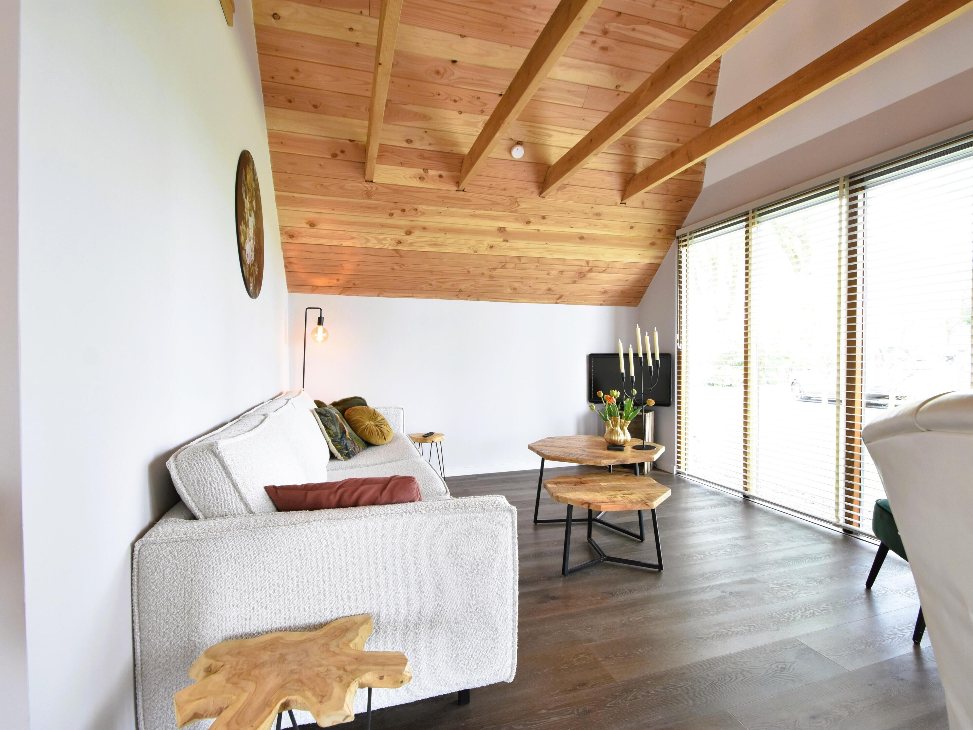 A modern living room with a high wooden ceiling, a grey sofa with cushions, a small wooden table with candles, and a large window with blinds.