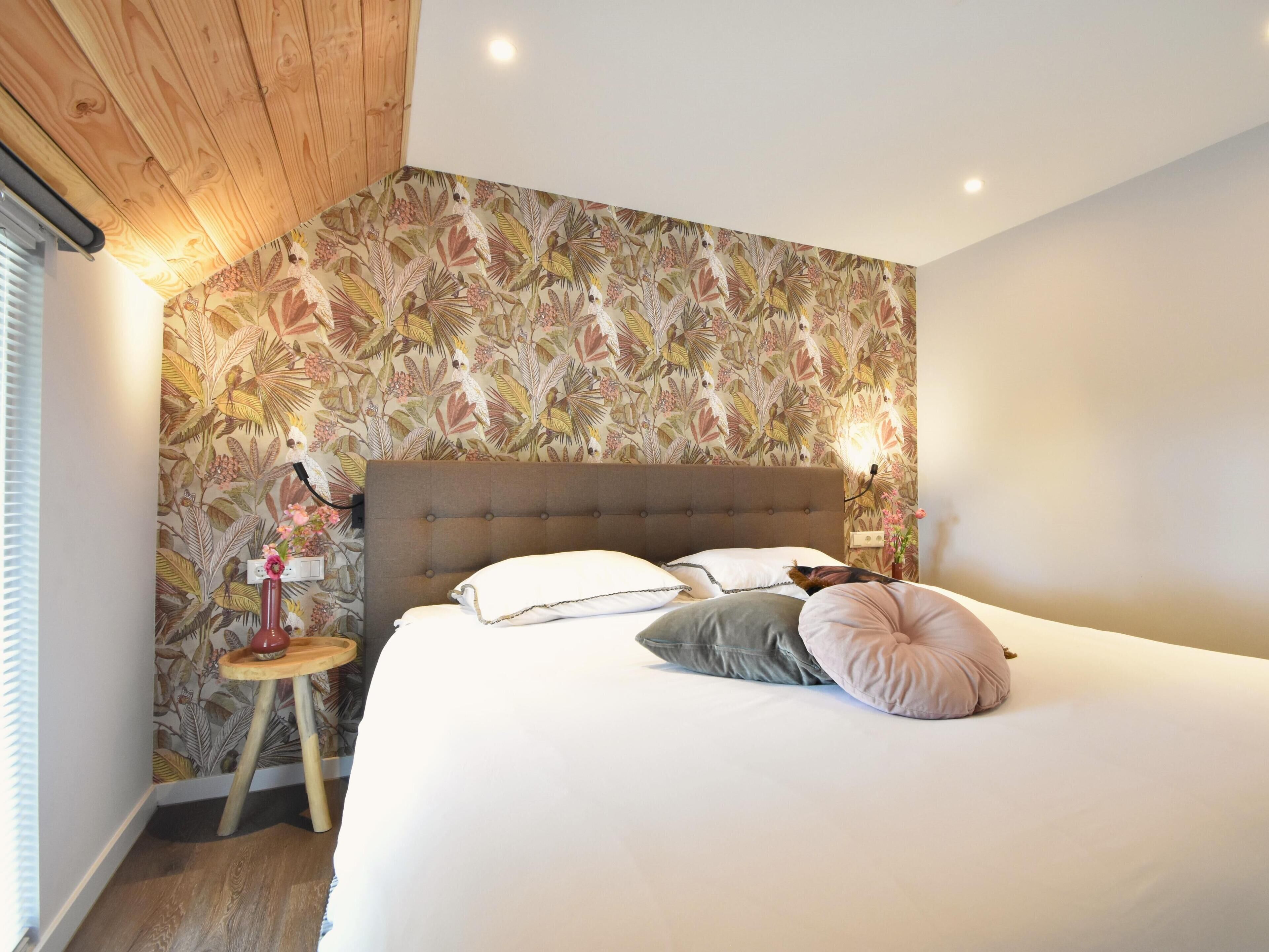Een slaapkamer met een groot bed, een houten hoofdbord, bloemenbehang, een nachtkastje met een lamp en een klein krukje.
