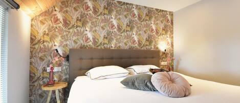 A bedroom with a large bed, a wooden headboard, a floral wallpaper, a bedside table with a lamp, and a small stool.