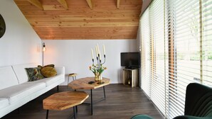 A modern living room with a wooden ceiling, a white sofa, a green armchair, a wooden coffee table, and a television on a stand.