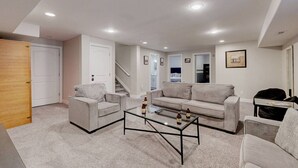 A modern living room with a sofa, armchair, and coffee table, a staircase leading to a bedroom, and a door to another room.