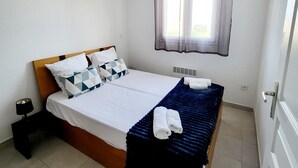 Una cama bien hecha con una colcha azul y toallas blancas, una lámpara de noche y una ventana con cortinas traslúcidas.
