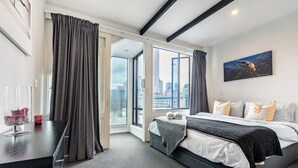 A modern hotel room with a large bed, a desk, and a view of the city through large windows. - Auckland Penthouse: SkyTower Views. Special Offer (Auckland)
