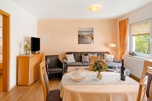 Une pièce comprenant un canapé, une table à manger avec un vase et des verres à vin, un téléviseur et une fenêtre avec des rideaux.