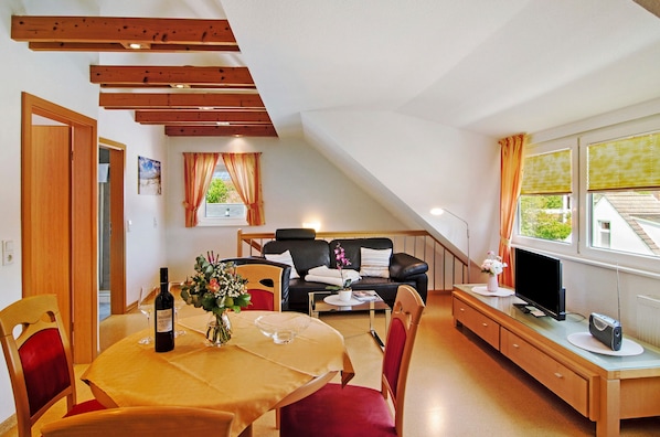 Ein Essbereich mit Tisch für zwei Personen, einer Couch, einem Fernseher und einem Fenster mit Vorhängen.