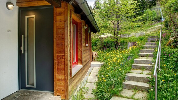 A wooden cabin with a dark door, a red window, and a stone staircase leading to a garden with wildflowers. - Holiday Home 'Berghütte Waldhäuser' with Mountain View, Private Terrace and Wi-Fi (Neuschönau)
