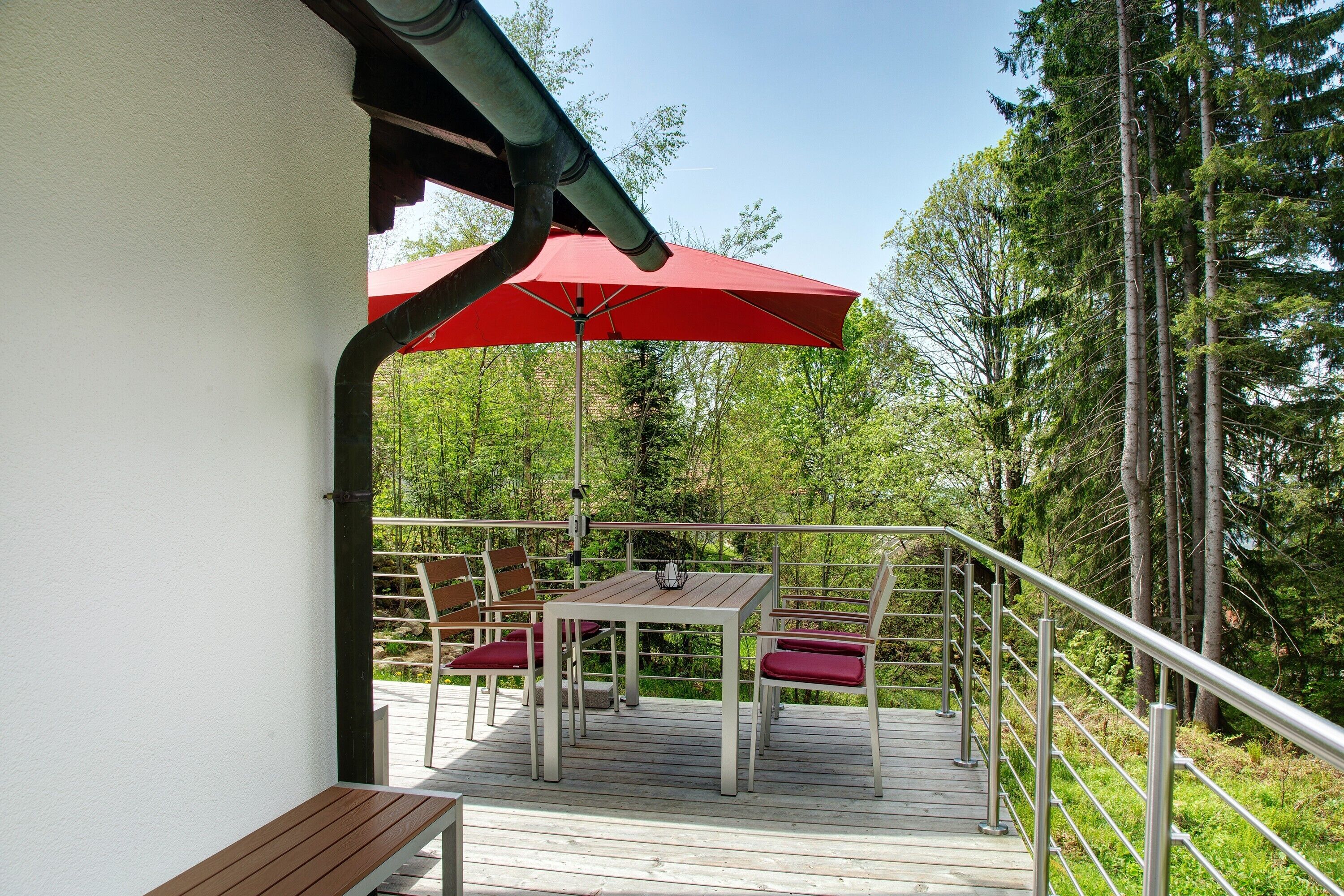 A deck with a table and chairs, a red umbrella, and a railing overlooking a forested area.