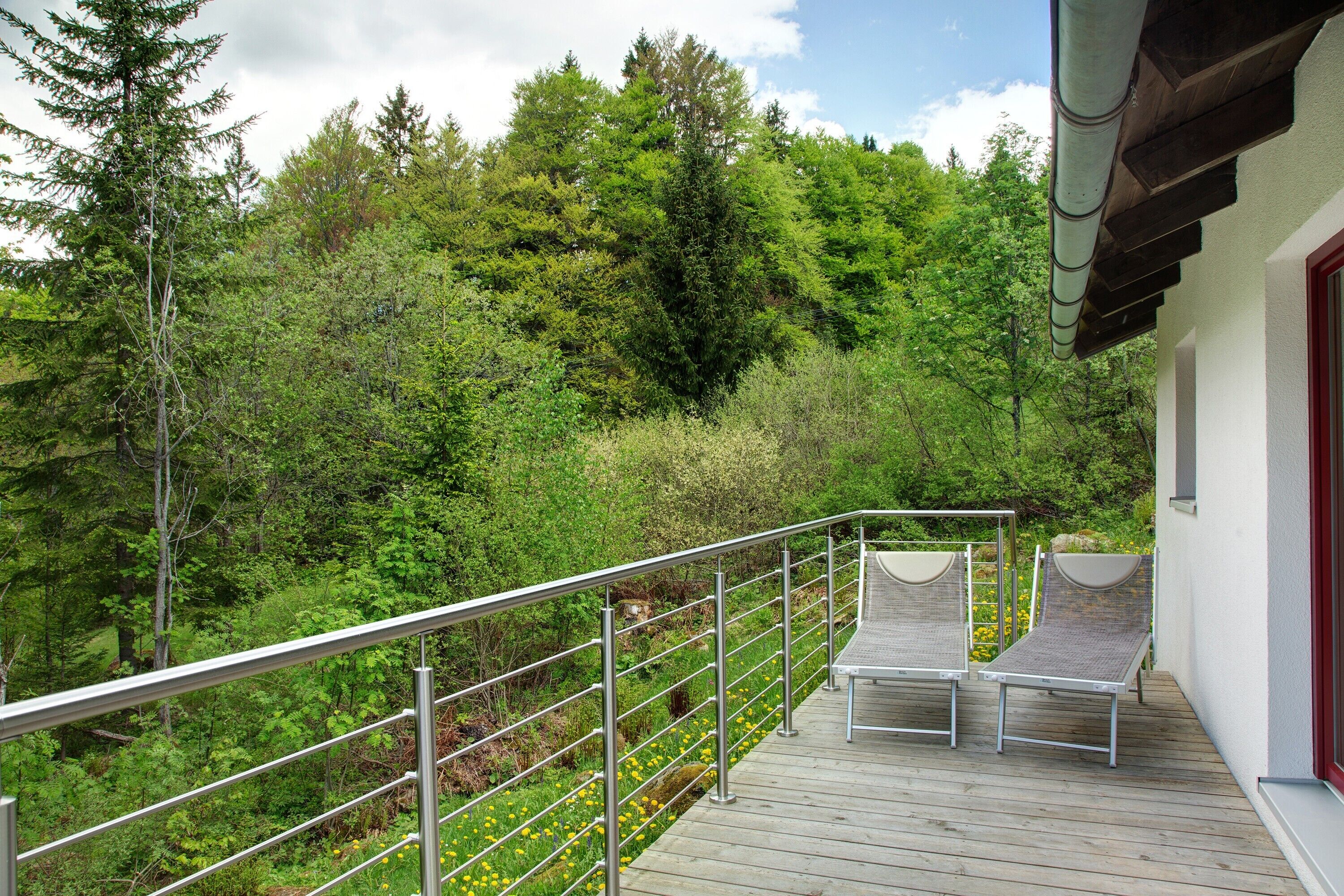 Une terrasse en bois avec des rampes en métal, deux chaises longues et une vue sur une colline boisée.