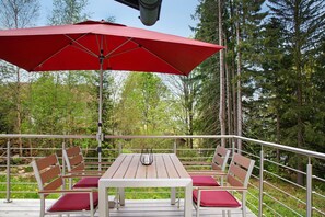 Un espace extérieur aménagé pour les repas, avec une table et des chaises, un parasol rouge et une vue sur les arbres.