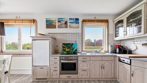 Une cuisine équipée d’un réfrigérateur, d’un four, d’un évier et de placards. Trois tableaux représentant des scènes de plage sont accrochés au mur.