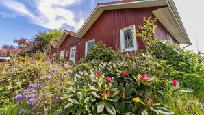 A red wooden house with a sloped roof, surrounded by lush greenery and flowering plants. - Holiday Apartment 'Erika' – Quiet Location with Large Garden near the Baltic Sea & Schlei (Saustrup)