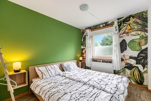 A bedroom with a bed, a nightstand with a lamp, a window with curtains, and a wall with a tropical leaf pattern. - Holiday Apartment 'Erika' – Quiet Location with Large Garden near the Baltic Sea & Schlei (Saustrup)