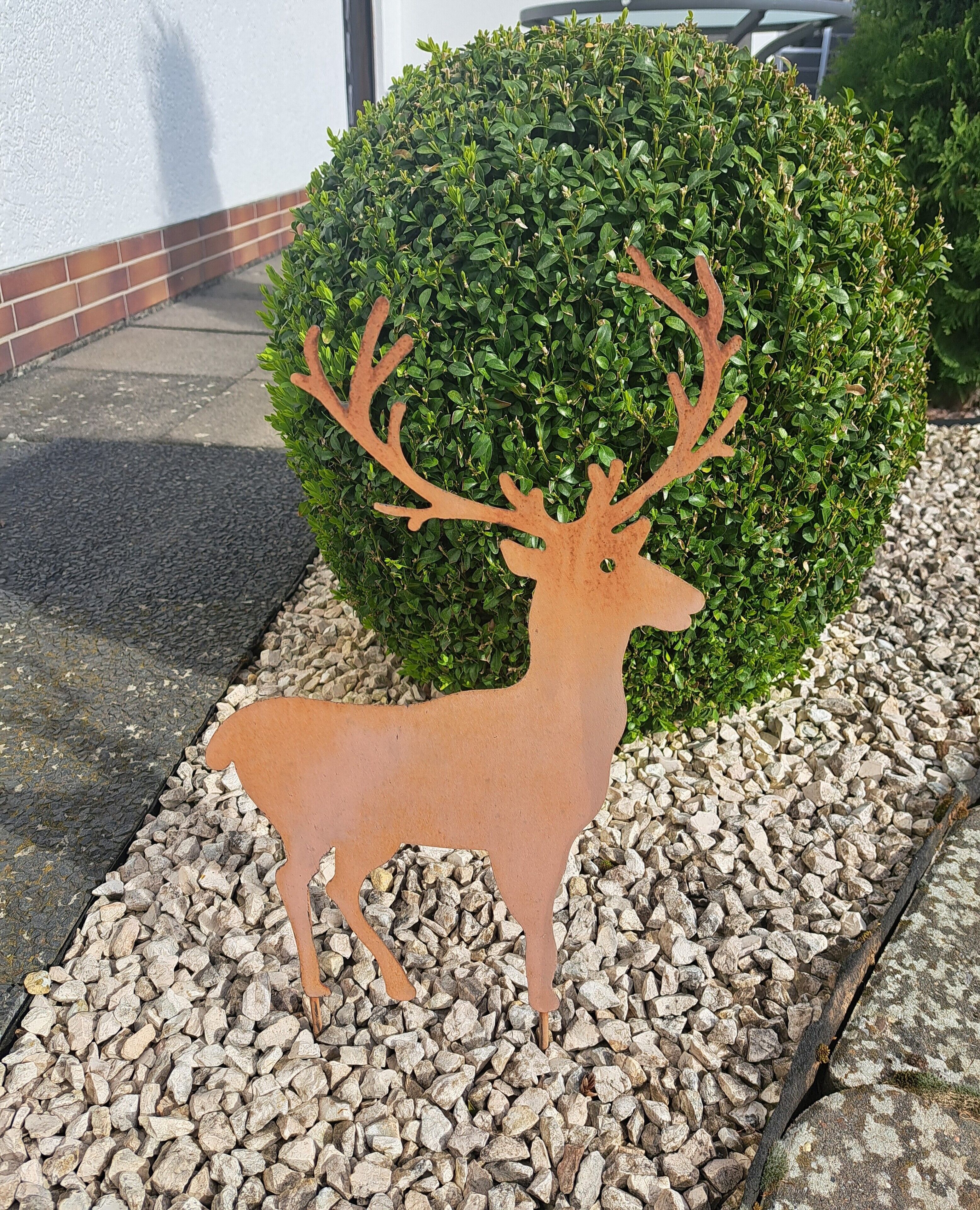 A metal deer sculpture in a landscaped area with gravel and a bush.