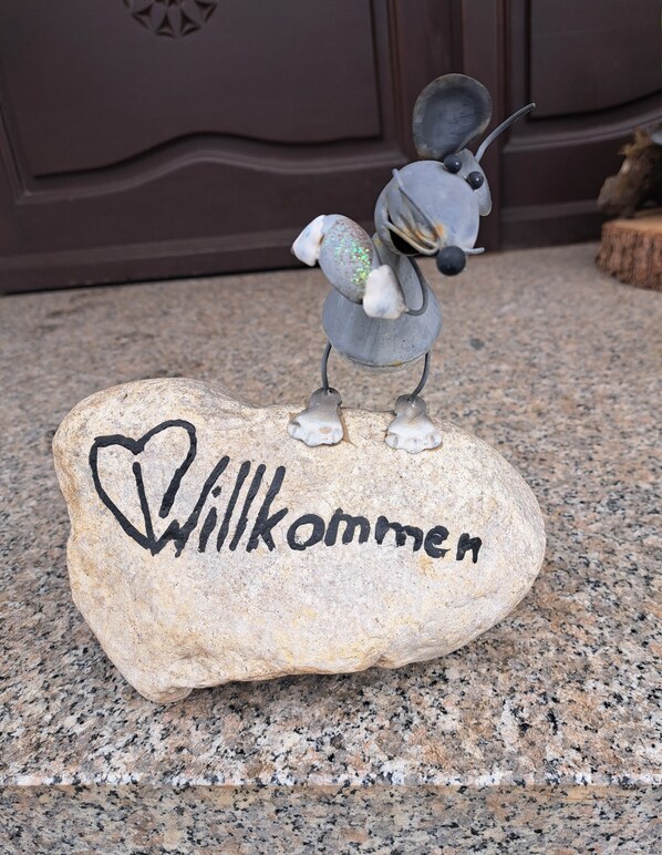 Een rots met 'Willkommen' en een hartsymbool, een beeldje van een persoon met een vogel, en een houten deur op de achtergrond.