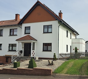 A two-story house with a front porch, a small garden, and a wooden fence. - Pension Andrea: Apartment 3 with Wi-Fi (Herbstein)