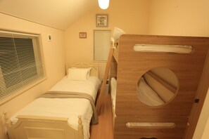 A compact bedroom with a single bed, a wooden bunk bed, a window with blinds, a bedside table, and a small framed picture on the wall. - Maccha House Nagano Zenko-ji (Nagano)