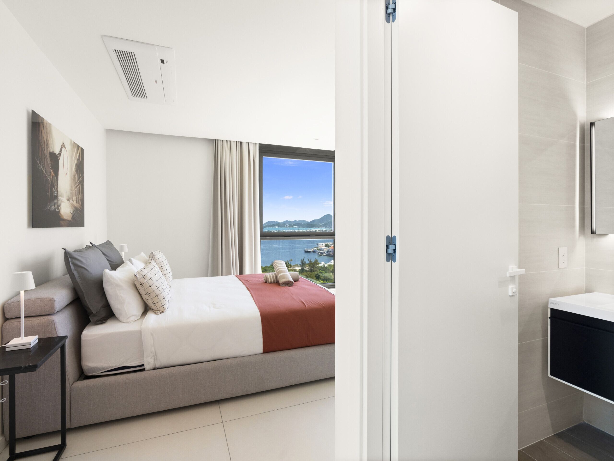A modern hotel room with a bed, a nightstand, a lamp, a window with a view of the sea, and a bathroom with a mirror and sink.