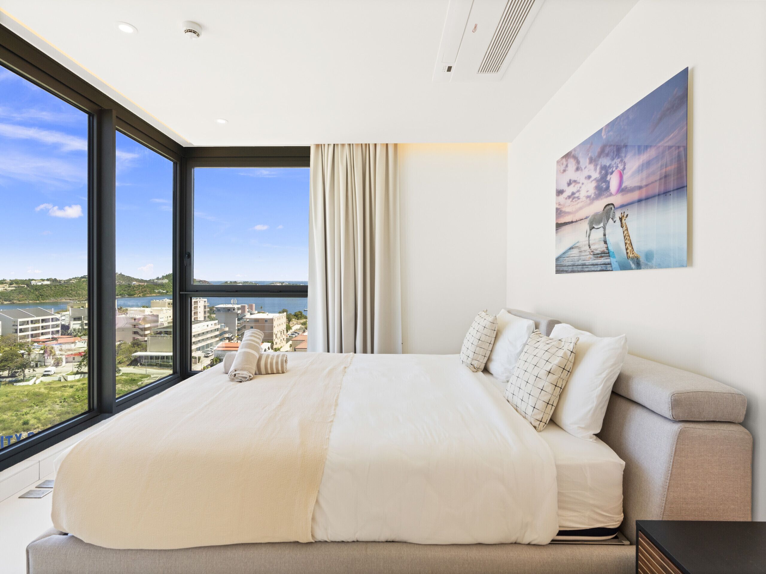 A modern hotel room with a large bed, a city view, and a painting on the wall.