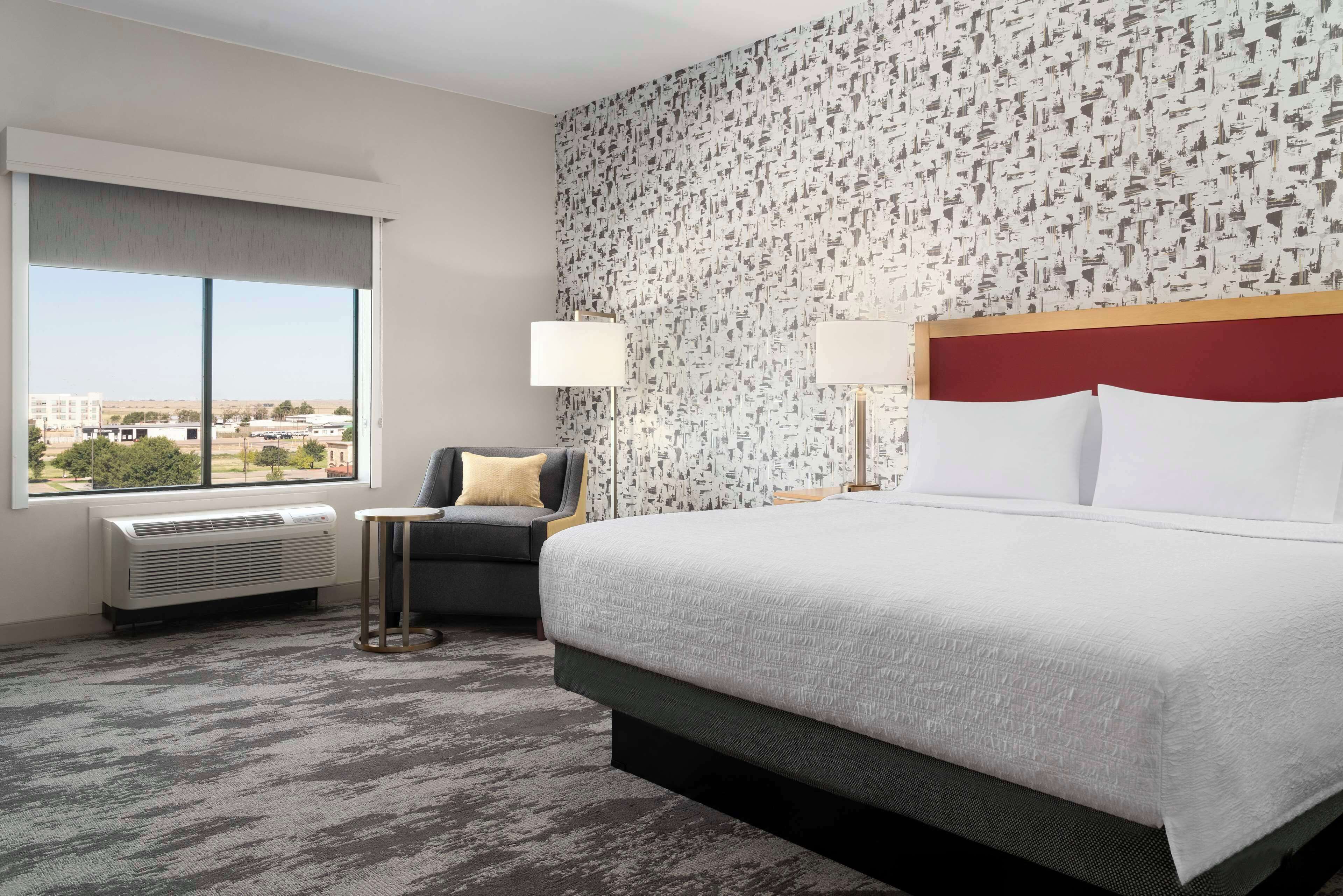 Habitación de hotel con una cama grande, una mesita pequeña, una silla, un ventanal con vistas y una pared con diseño.