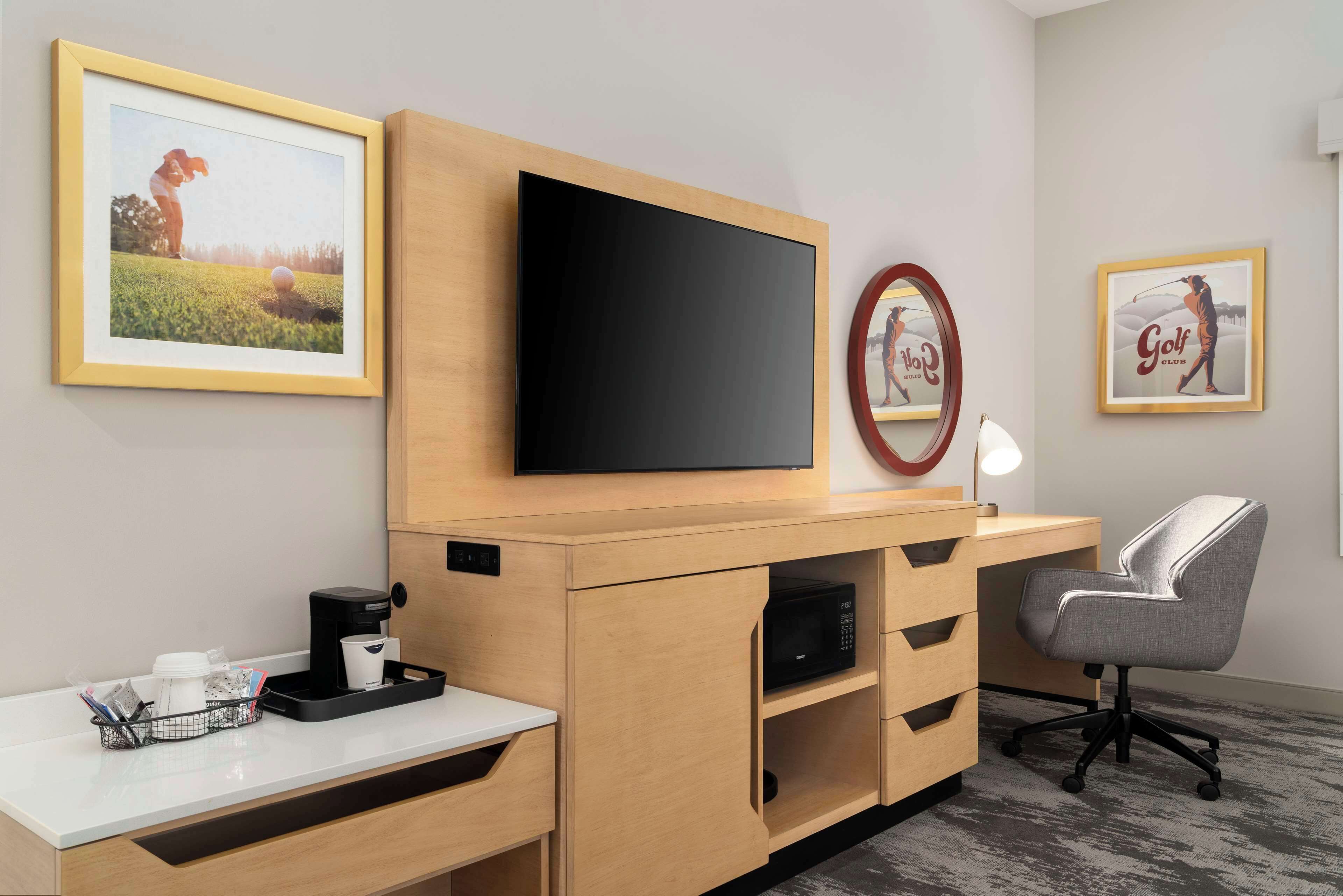 A modern hotel room with a wooden desk, a flat-screen TV, a grey office chair, and framed golf-themed artwork on the walls.