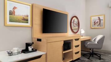 A modern hotel room with a wooden desk, a flat-screen TV, a grey office chair, and framed golf-themed artwork on the walls.