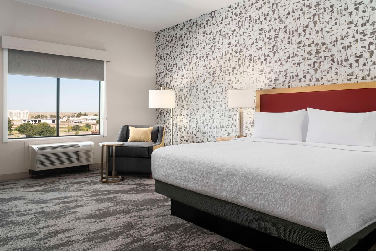 A hotel room with a large bed, a small sofa, a side table, a lamp, and a view of the city through a window.