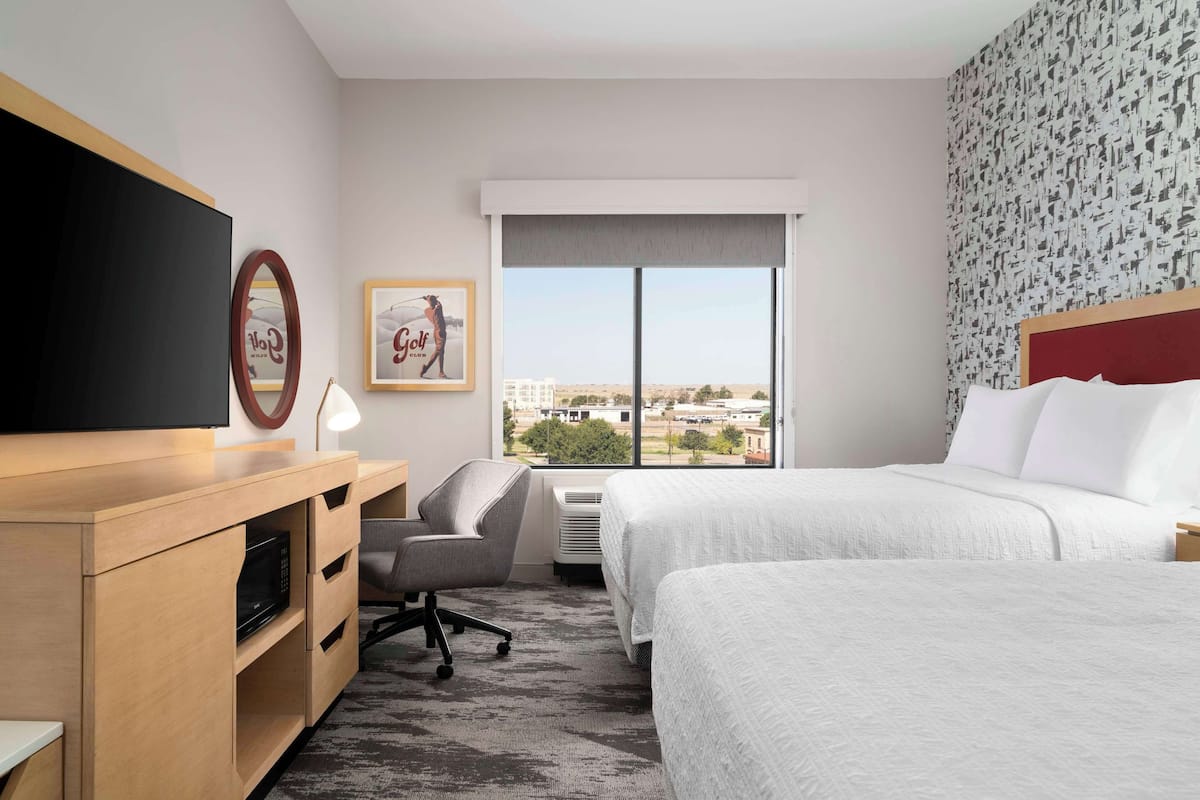 A hotel room with a bed, a desk with a TV, a chair, a framed picture, and a window with a city view.