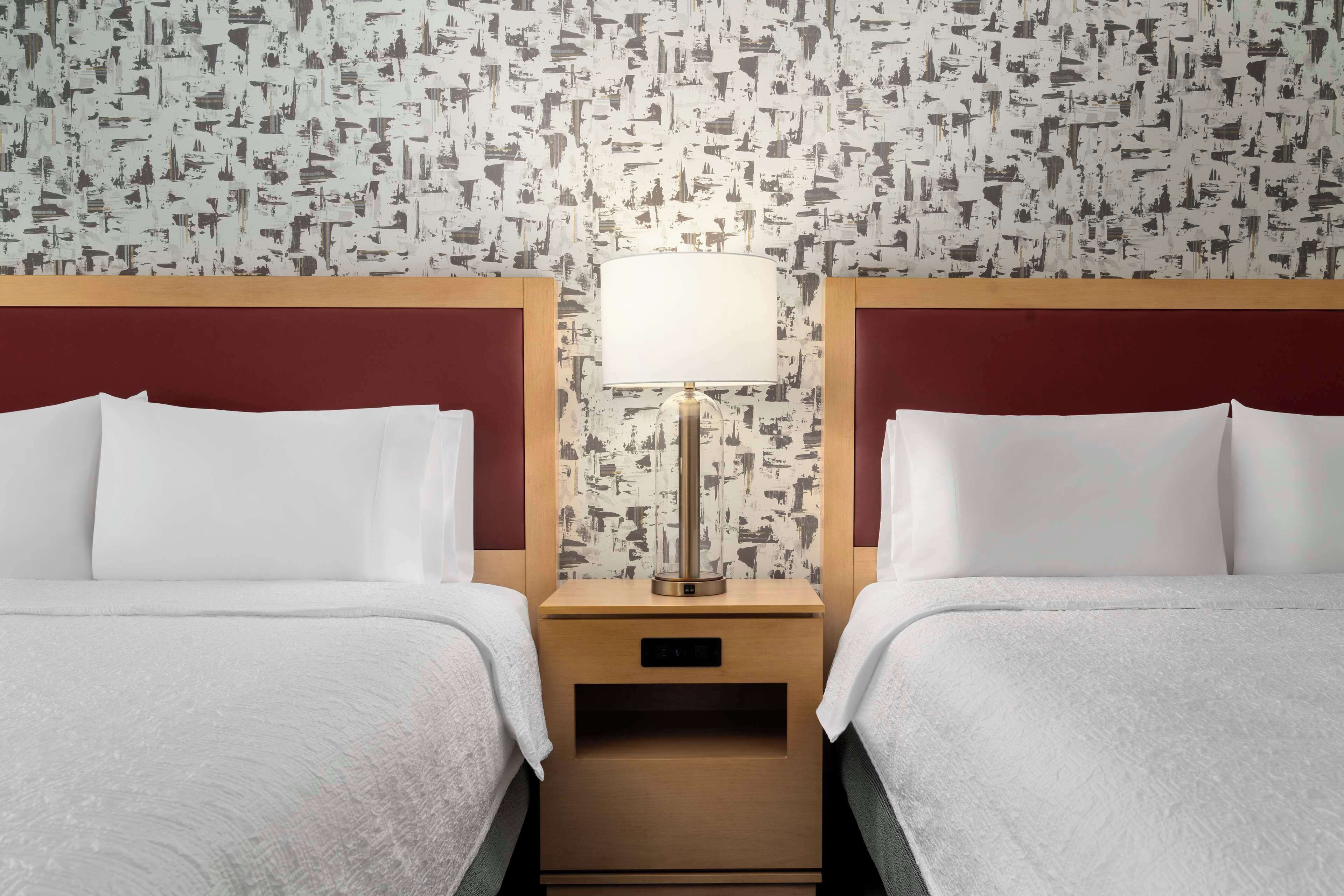 A hotel room with two beds, a nightstand with a lamp, and a patterned wallpaper.