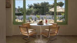 A dining area with a round table and four wicker chairs, a window with green frames, and a view of a garden with trees and a white umbrella. - Kallisti Stay (Spata-Artemida)