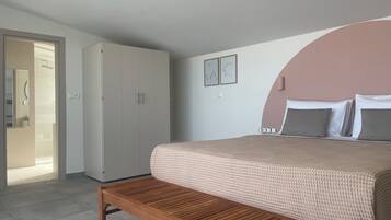 Un dormitorio con cama, un banco de madera, un armario y un baño visible a través de una puerta abierta.