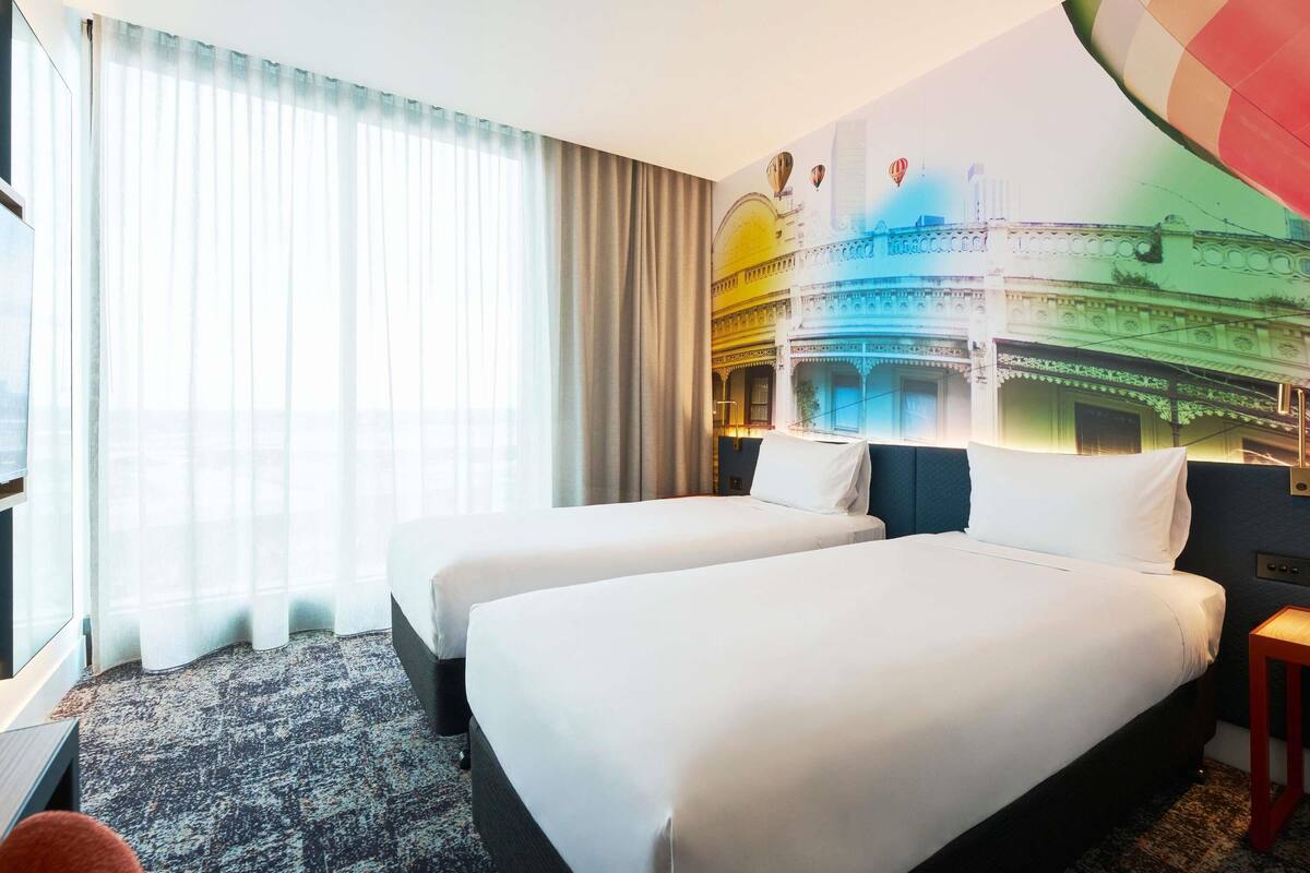 Een hotelkamer met twee bedden, een groot raam met dunne gordijnen, een vloerbedekking en een kleurrijke muurschildering.