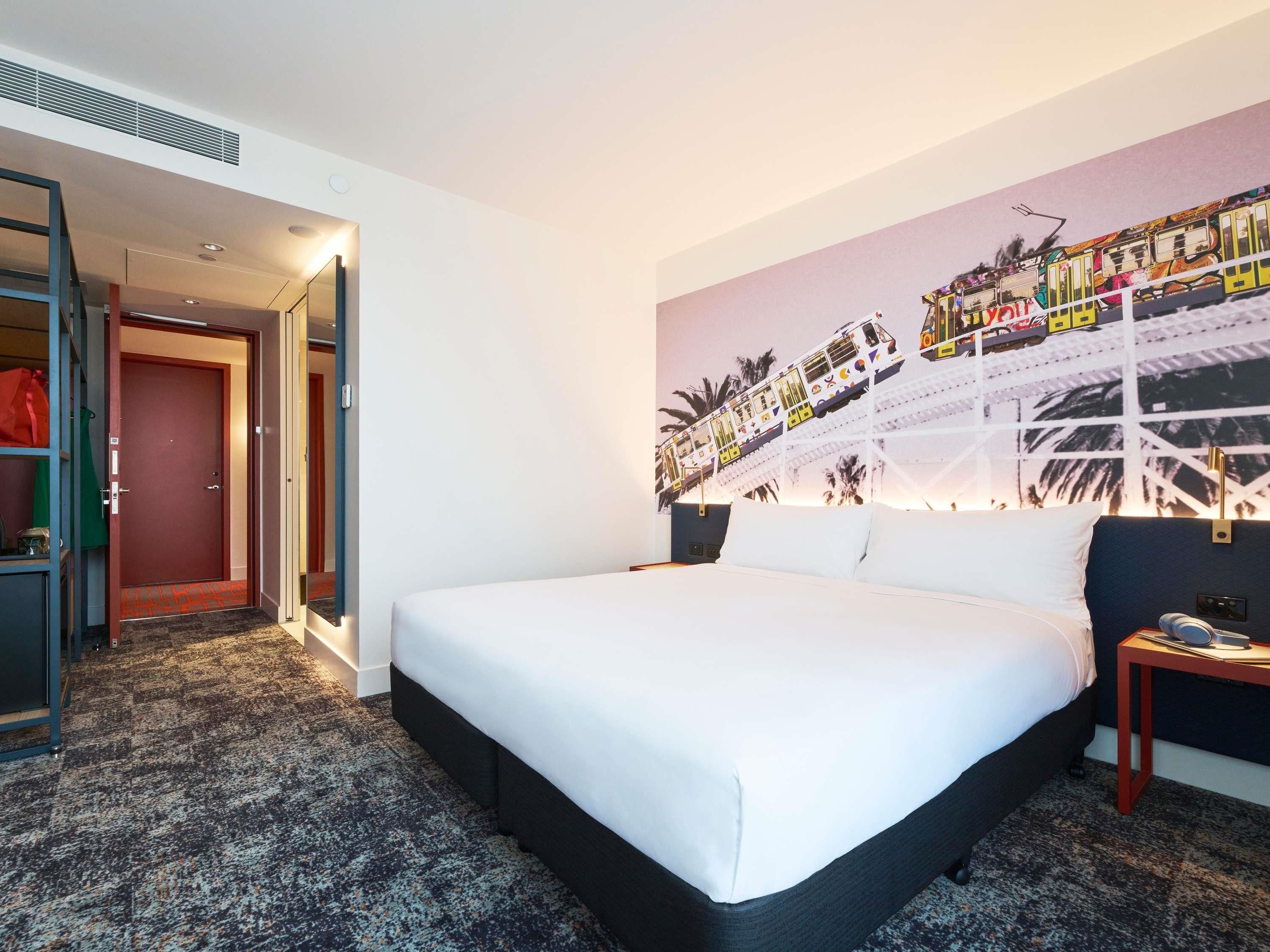 Ein Hotelzimmer mit einem großen Bett, einem Nachttisch, einem an der Wand befestigten Fernseher und einer Tür zu einem Flur.