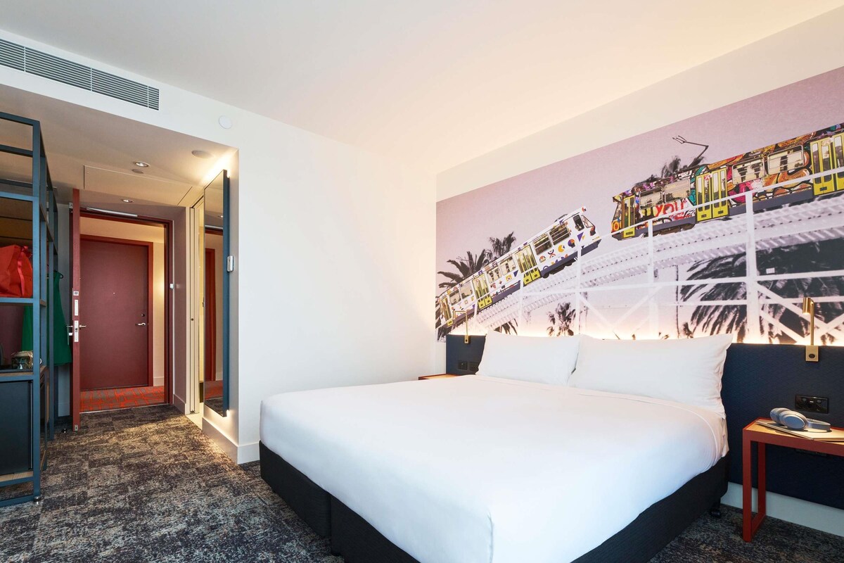 Une chambre d’hôtel avec un grand lit, une table de chevet, un téléviseur fixé au mur et une porte donnant sur un couloir.