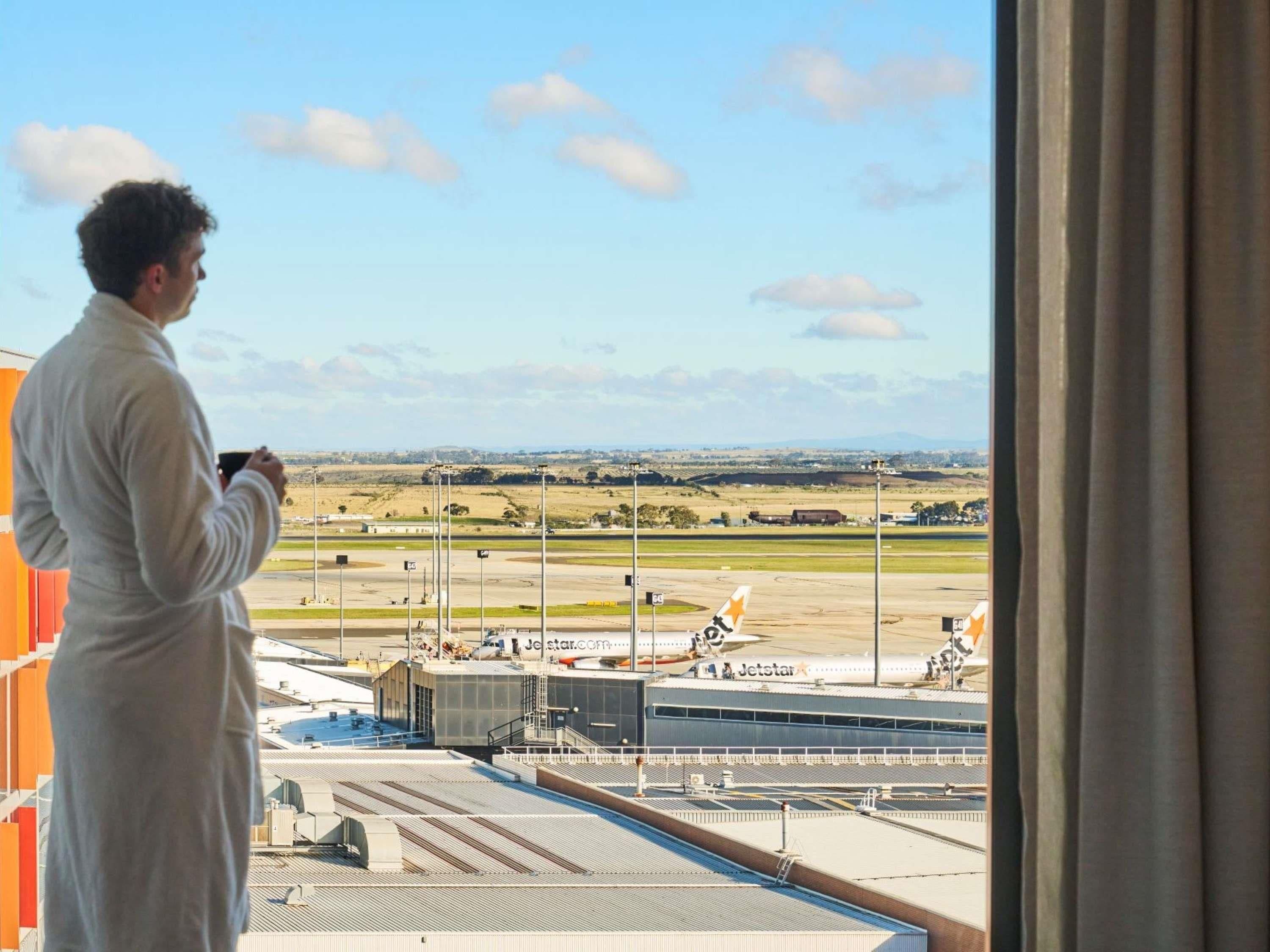 Una persona vestida con bata de baño de pie junto a una ventana con vistas a un aeropuerto, con pista y edificios visibles.