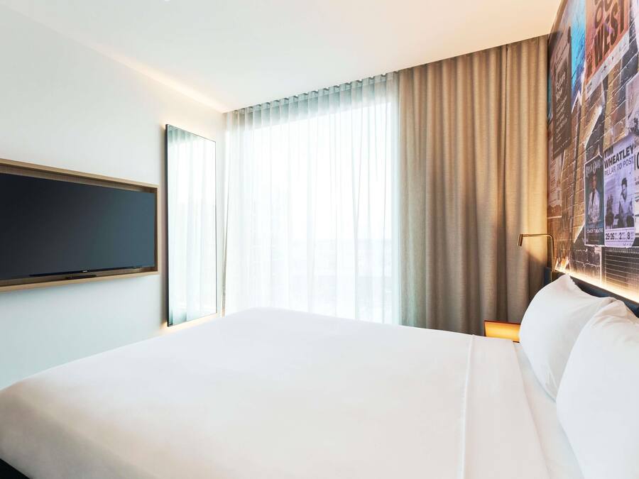 Una habitación de hotel con una cama grande, un televisor de pantalla plana montado en la pared, un ventanal con cortinas translúcidas y una pared con un cartel.