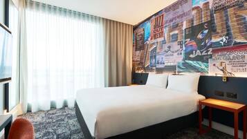 A hotel room with a large bed, a desk, a chair, and a wall decorated with event posters.
