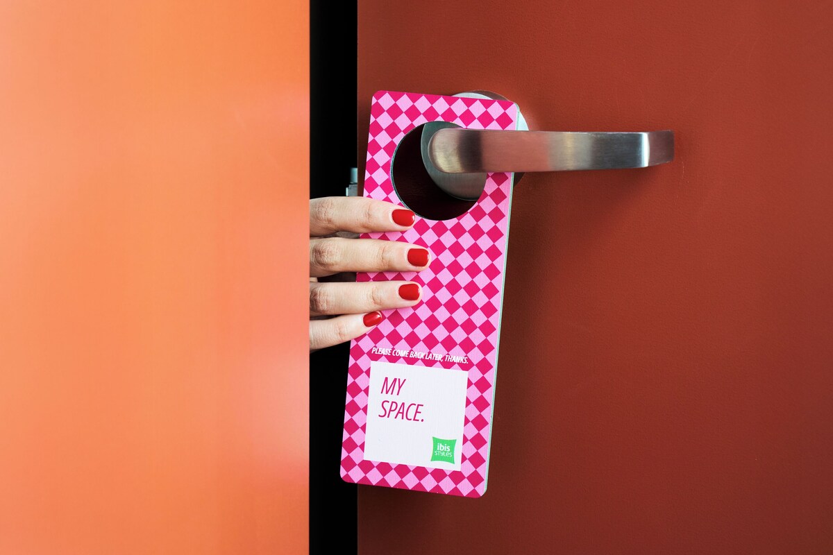 Une main tenant la poignée d’une porte, à laquelle est attachée une carte à carreaux roses et blancs sur laquelle on peut lire le texte « MY SPACE ».