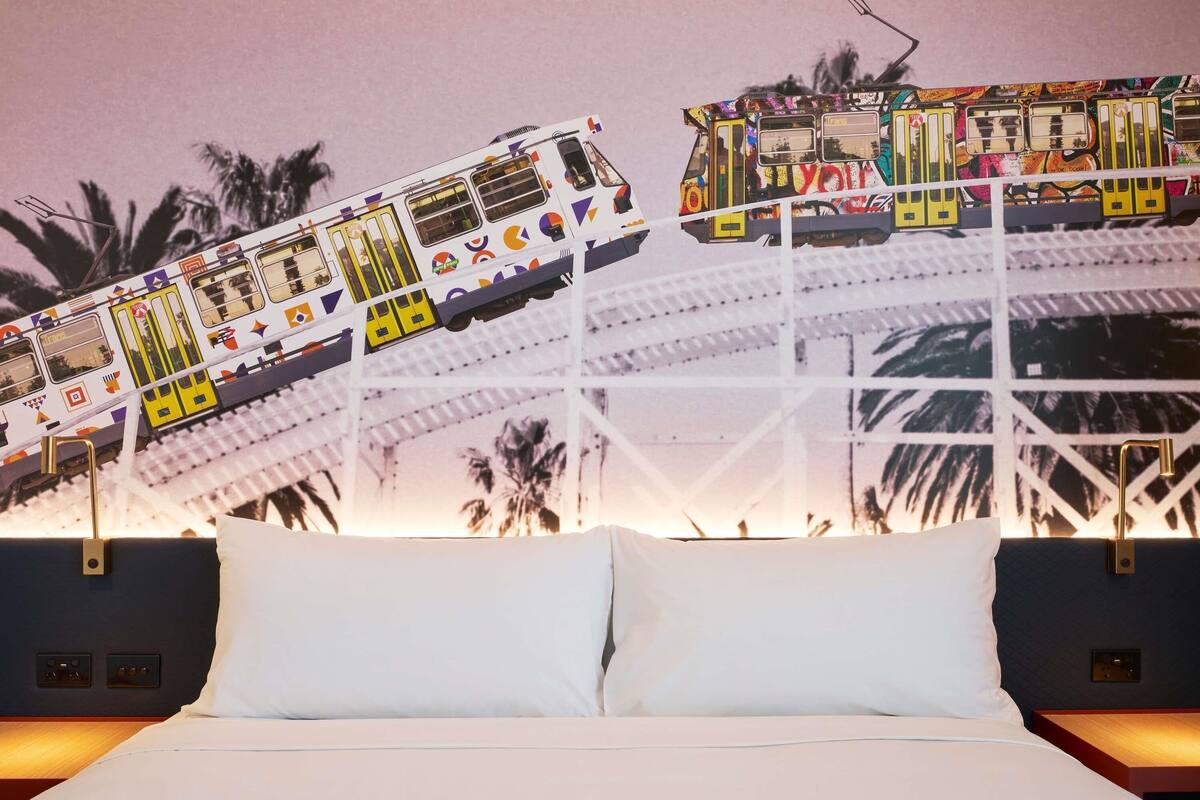 Een bed met witte lakens, een nachtkastje en een muur met een kleurrijk treinmuurdecoratie.
