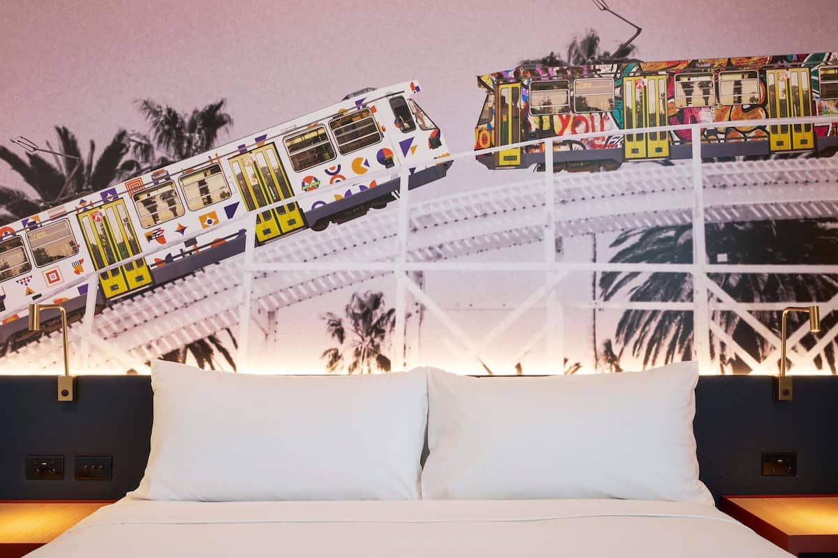 Un lit avec du linge de lit blanc, une table de chevet et un mur orné d’une fresque représentant un train coloré.