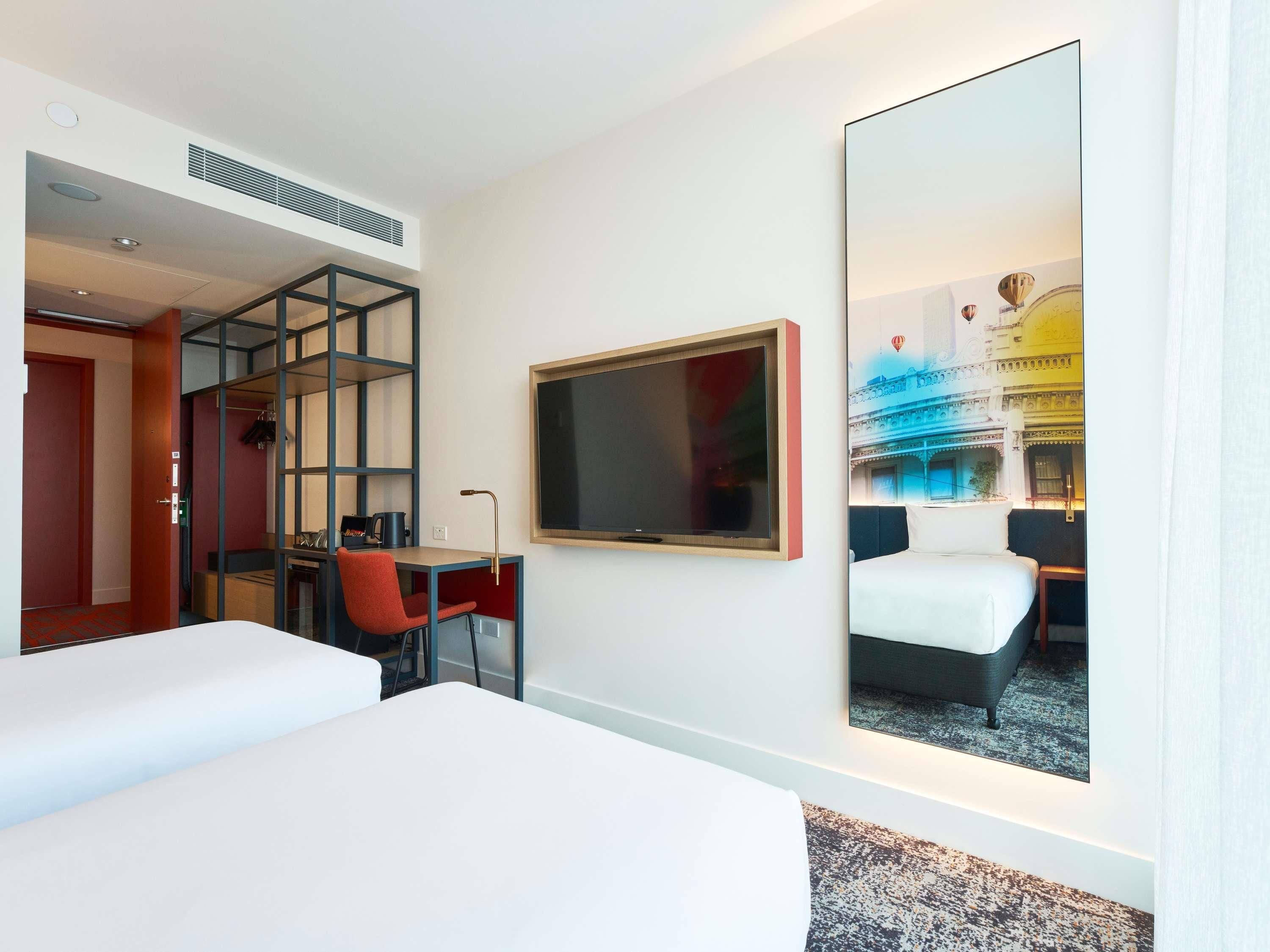 A hotel room with a bed, a desk with a chair, a bookshelf, and a large mirror reflecting a room with a bed and a decorative wall.
