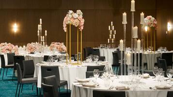 Un salón de banquetes con mesas redondas dispuestas para comer, cada una con mantel blanco, copas de cristal transparente y velas.